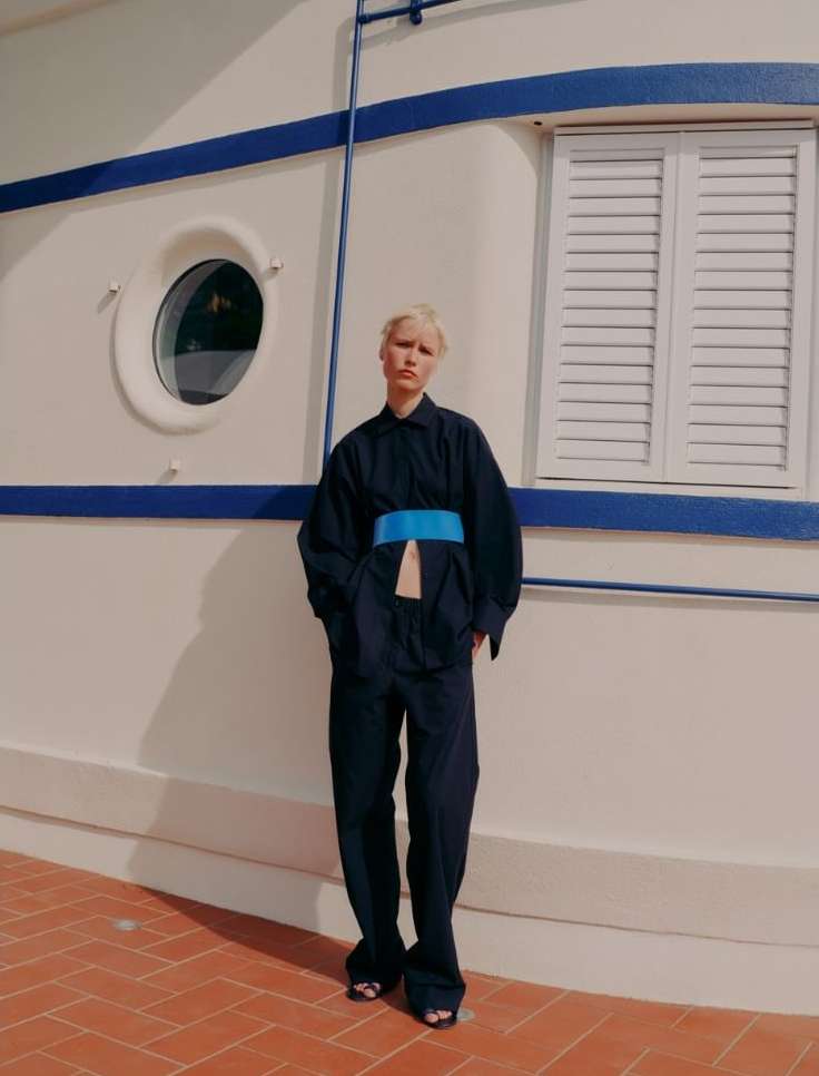 A short-haired model in a dark jumpsuit with a bright blue belt, poses in front of a white building with blue trim and shuttered window.