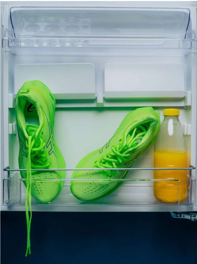 Inside a refrigerator, two neon green sneakers are placed next to a bottle of orange juice with a yellow cap on a shelf.