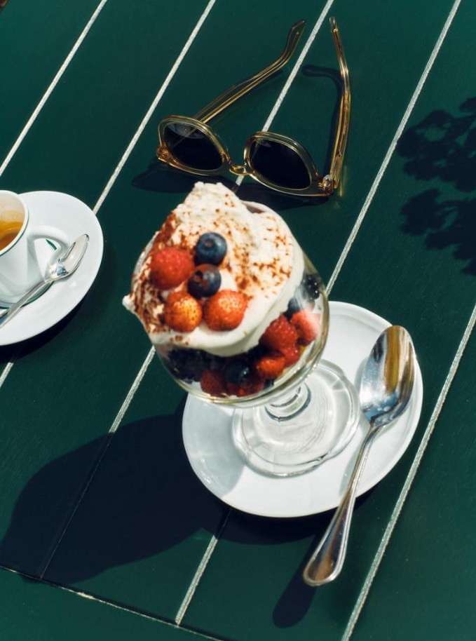 Overhead shot of a parfait with berries and whipped cream, sunglasses, and an espresso on a green striped table.