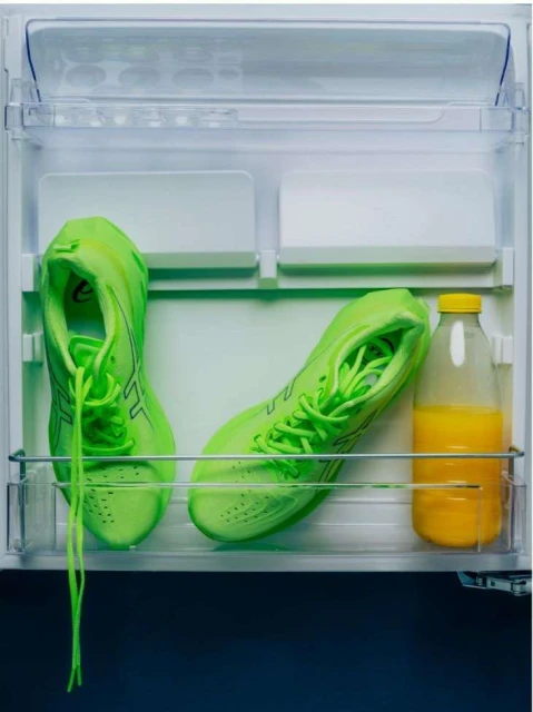 A pair of bright green running shoes sit inside a refrigerator on a shelf next to a plastic bottle of orange juice.