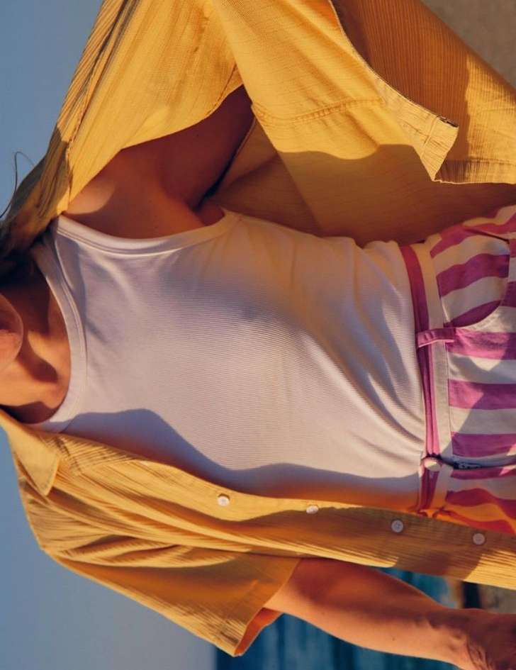 A person wearing a white tank top, pink striped shorts, and a yellow button-down shirt is partially visible. Sky in the background.