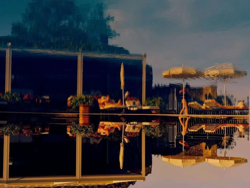 Outdoor patio reflected in rippled water, showing architecture, parasols, lounge chairs, and lush plants creating a mirrored, surreal scene.