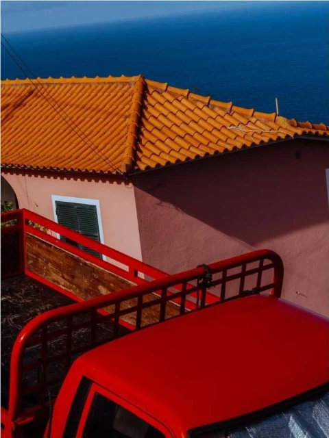 Red pickup truck parked next to a pink building with a terracotta tile roof with an ocean view in the background on a sunny day.