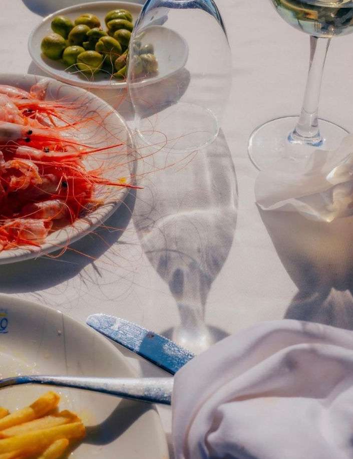 Overhead shot of a table with shrimp, olives, wine glasses, fries, silverware, and a white tablecloth in bright, summery light.