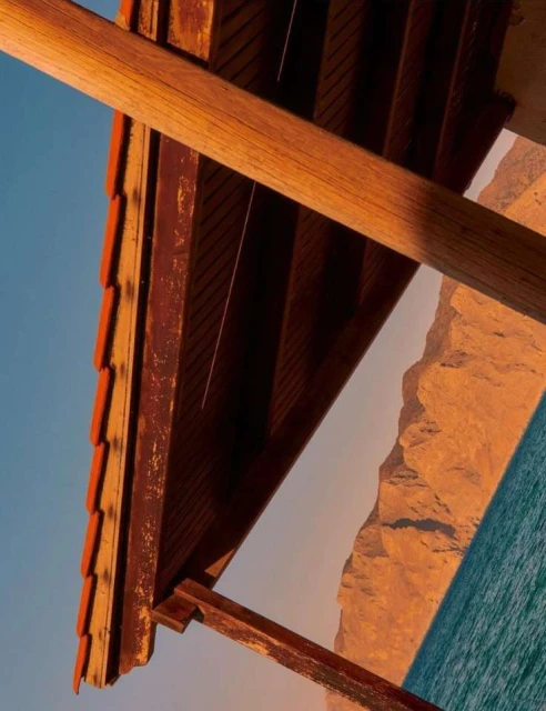 Angled view of a wooden structure with terracotta-colored tiles overlooking the sea and a mountain, captured during daytime.