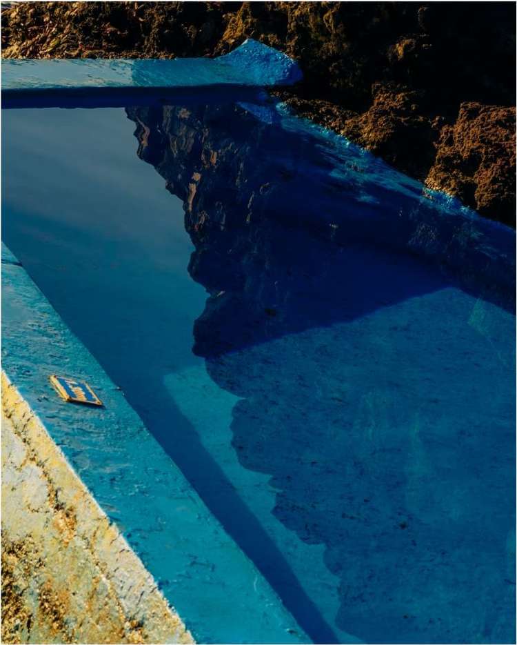 High-angle shot of a bright blue swimming pool being constructed, showing brown dirt and concrete with the reflection of the sky in the water.
