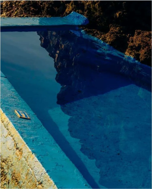 High-angle shot of a bright blue swimming pool being constructed, showing brown dirt and concrete with the reflection of the sky in the water.