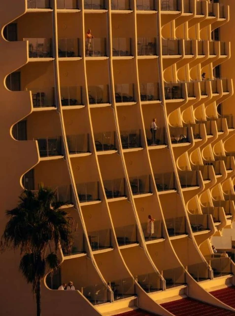 Exterior shot of modern, multi-story hotel featuring curved balconies and warm golden light, with people visible on balconies.