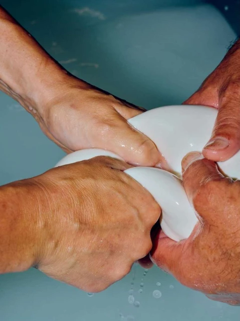 Close-up of two pairs of hands kneading and stretching a ball of mozzarella cheese in a basin of light-blue tinted water in an indoor setting.