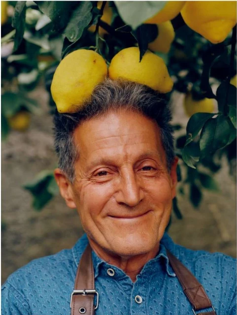 Elderly man smiles with lemons on his head in an outdoor setting. He is wearing a blue shirt with a brown apron and the background shows green leaves.