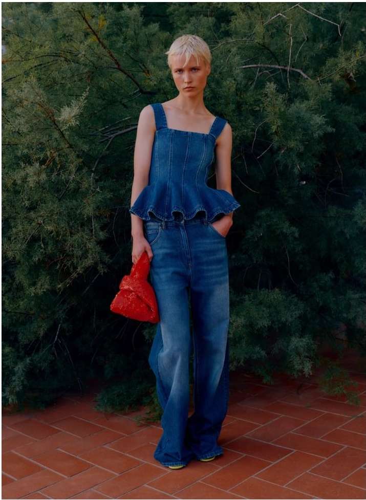 Full-length shot of a female model in a denim outfit with an orange purse, posing in front of a dark green shrubbery background on a brick surface.