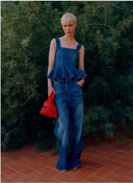 Full-length shot of a female model in a denim outfit with an orange purse, posing in front of a dark green shrubbery background on a brick surface.