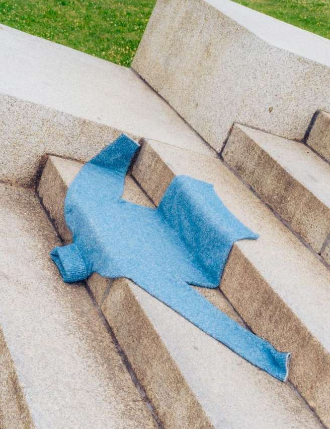 Light blue knitted sweater is laying on concrete steps, outdoors on a sunny day. The steps are multi-tiered surrounded by green grass.
