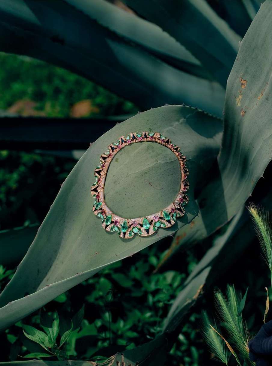 Close-up of a gold necklace with emeralds and rubies, lying on a large agave plant leaf, set against a green leafy background.