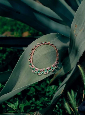 Close-up of a gold necklace with emeralds and rubies, lying on a large agave plant leaf, set against a green leafy background.