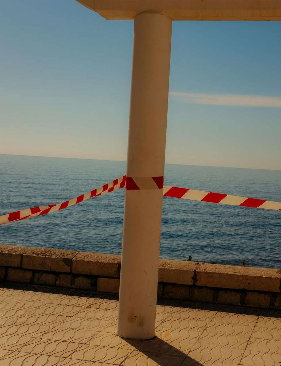 Concrete pillar surrounded by red and white striped barrier tape against a backdrop of the ocean and blue sky, creating a sense of closure.