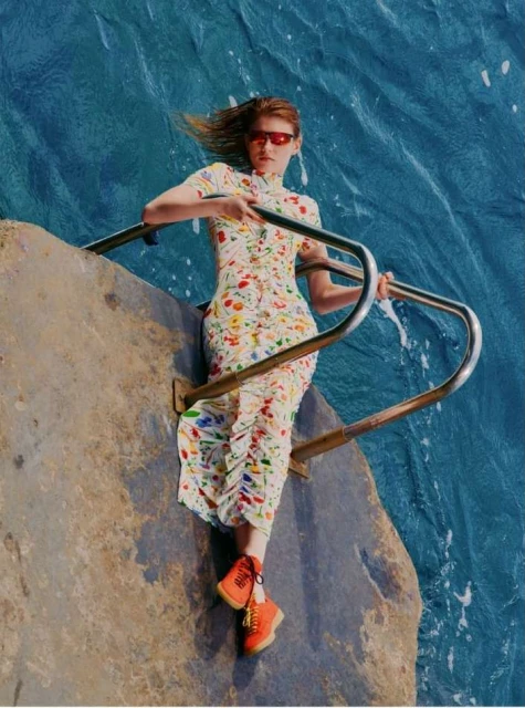 A woman in a floral dress and red sunglasses leans on a railing by the ocean. She is wearing orange shoes in a fashion style editorial shot.