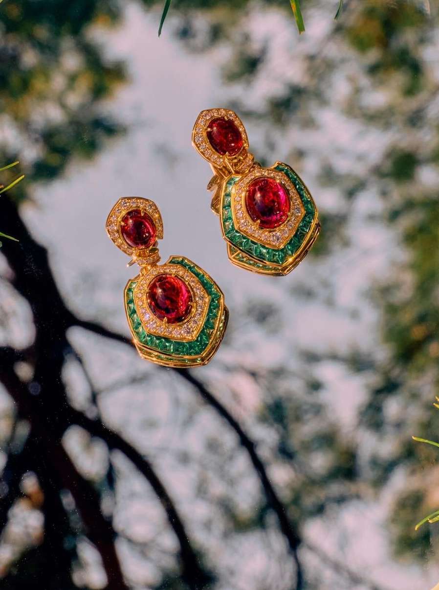 Luxurious gold drop earrings with red gemstones and green accents showcased against a blurred natural backdrop, jewelry advertisement.