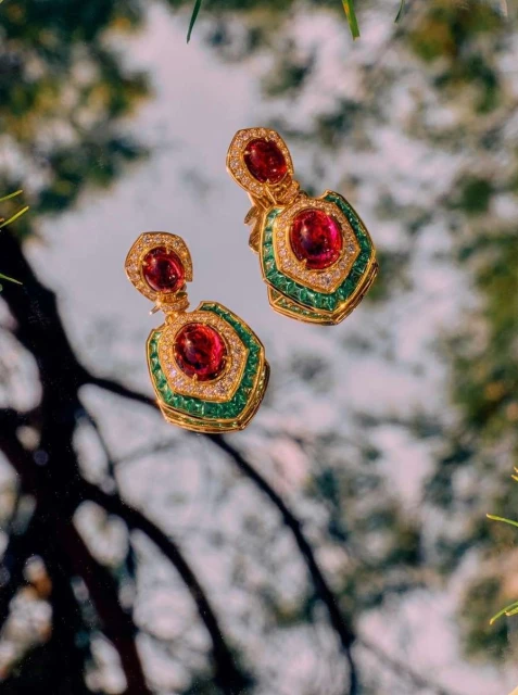 Luxurious gold drop earrings with red gemstones and green accents showcased against a blurred natural backdrop, jewelry advertisement.
