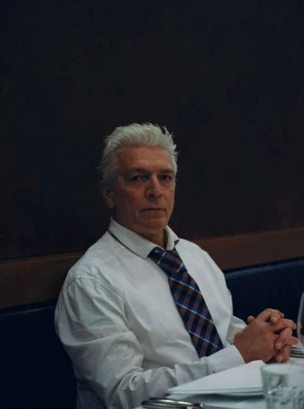 Older man with white hair wearing a white dress shirt and plaid tie seated at a table, photographed by Maximilian Virgili.