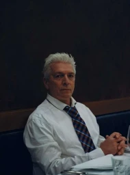 Older man with white hair wearing a white dress shirt and plaid tie seated at a table, photographed by Maximilian Virgili.