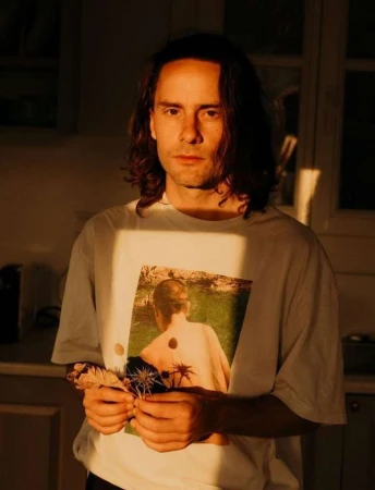 Portrait of a man with long dark hair wearing a white t-shirt with a graphic print, holding dried leaves under warm sunlight.
