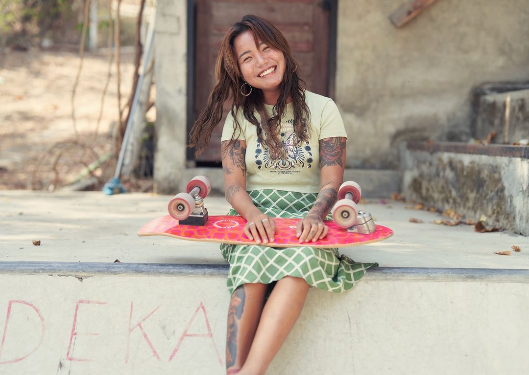 Smiling woman with tattoos and skateboard sitting on a concrete step, wearing a t-shirt and patterned skirt in an outdoor setting.