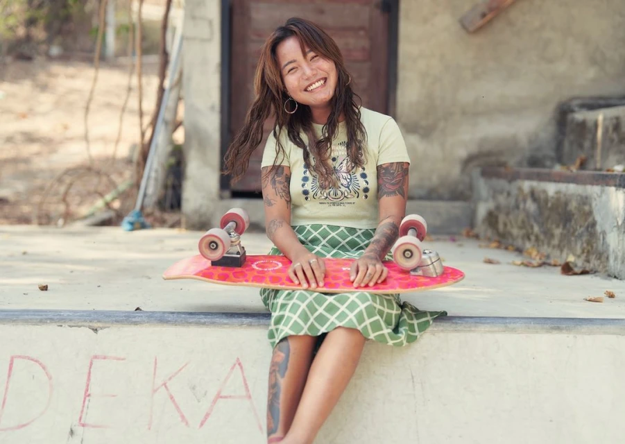 Smiling woman with tattoos and skateboard sitting on a concrete step, wearing a t-shirt and patterned skirt in an outdoor setting.
