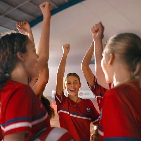 A group of young female athletes in red uniforms celebrate a win indoors, raising their arms with joyous expressions and smiles.