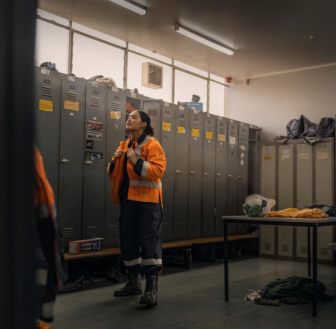 Woman in an orange safety vest stands in a locker room, preparing for work. Industrial setting, safety gear, and workwear are visible.