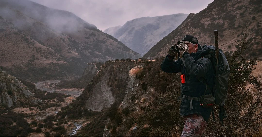 Adult male uses binoculars in a mountain valley. He wears a backpack with a rifle and camouflage pants. Cloudy day and low angle shot.