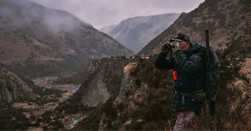 Adult male uses binoculars in a mountain valley. He wears a backpack with a rifle and camouflage pants. Cloudy day and low angle shot.