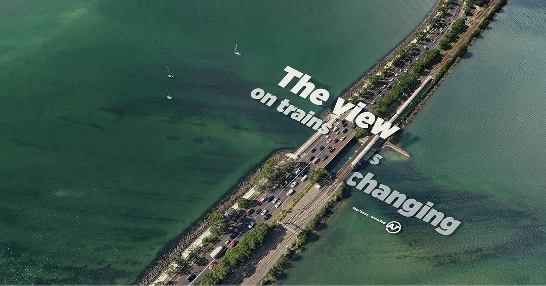 Aerial view of a bridge with car and train traffic over a green body of water, featuring sailboats and promotional text overlay.