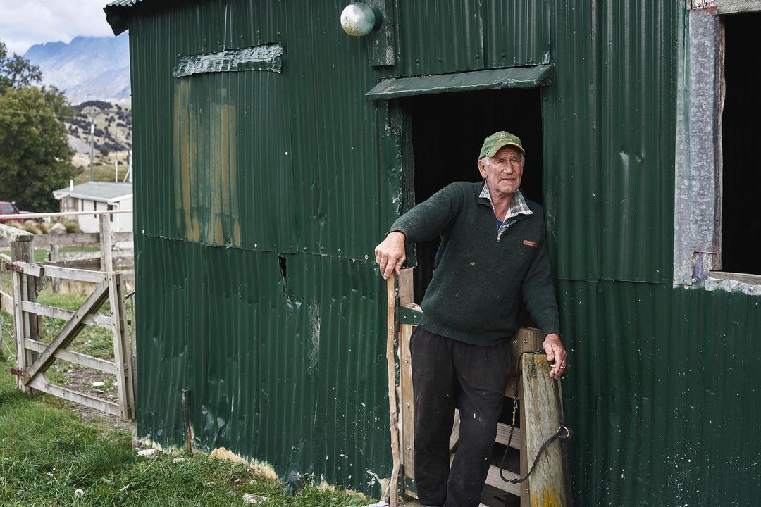 Elderly man in green sweater stands in doorway of corrugated metal building in rural setting with mountains in the background.