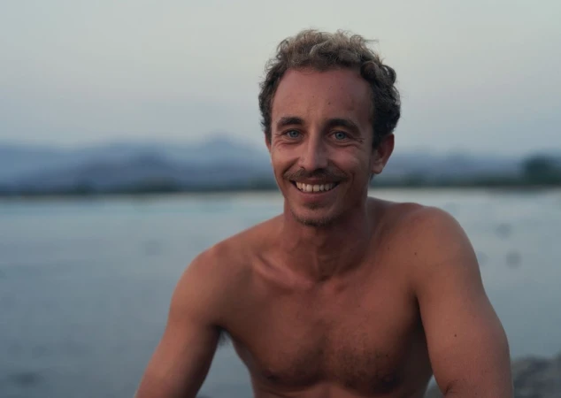 Smiling shirtless man with a mustache outdoors against a soft-focus landscape background.