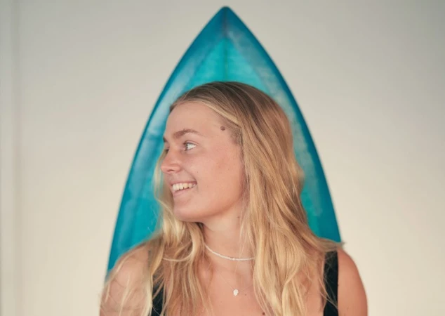 Young blond woman smiling in profile in front of a blue surfboard against a plain white wall background
