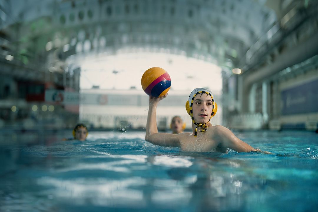 Teen boy throwing a water polo ball in an indoor swimming pool, wearing a water polo cap, with other players in the background.