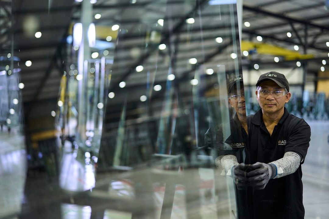 Worker in a factory holding a large glass panel in front of rows of glass and blurred lights. The man wears a cap and glasses.