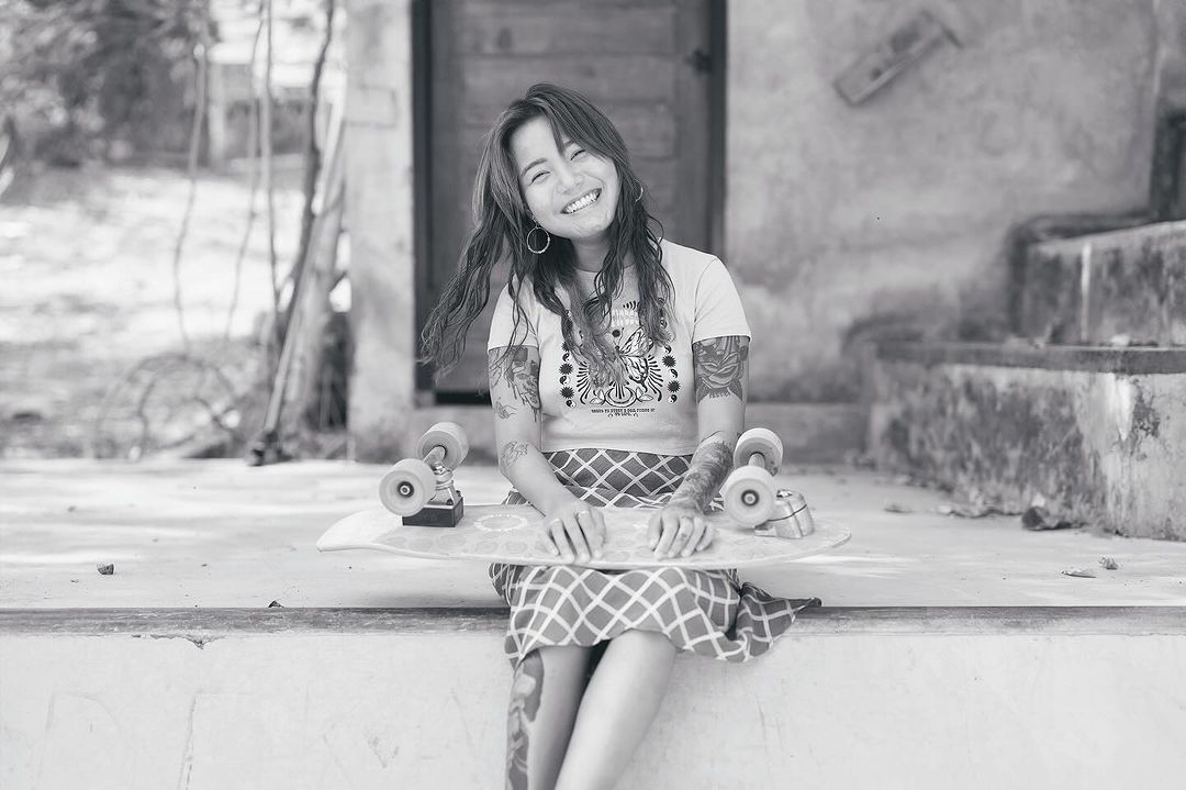 Smiling woman with arm tattoos sitting on a concrete step holding a skateboard with a doorway and greenery in the background.