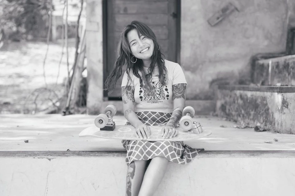Smiling woman with arm tattoos sitting on a concrete step holding a skateboard with a doorway and greenery in the background.