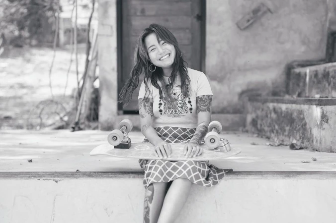 Smiling woman with arm tattoos sitting on a concrete step holding a skateboard with a doorway and greenery in the background.