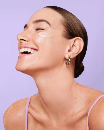 Close-up of a smiling woman with her eyes closed, wearing a cream dot on her cheek and silver hoop earrings against a purple background.