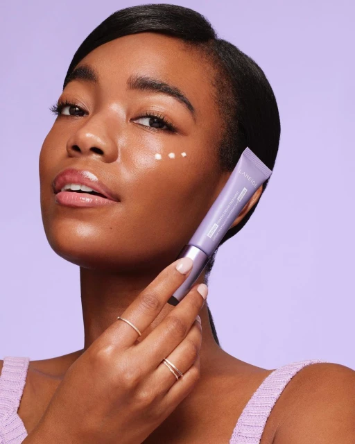 Close-up product shot of a woman applying skincare cream to her face. Lilac background, cream tube, gold rings on fingers.