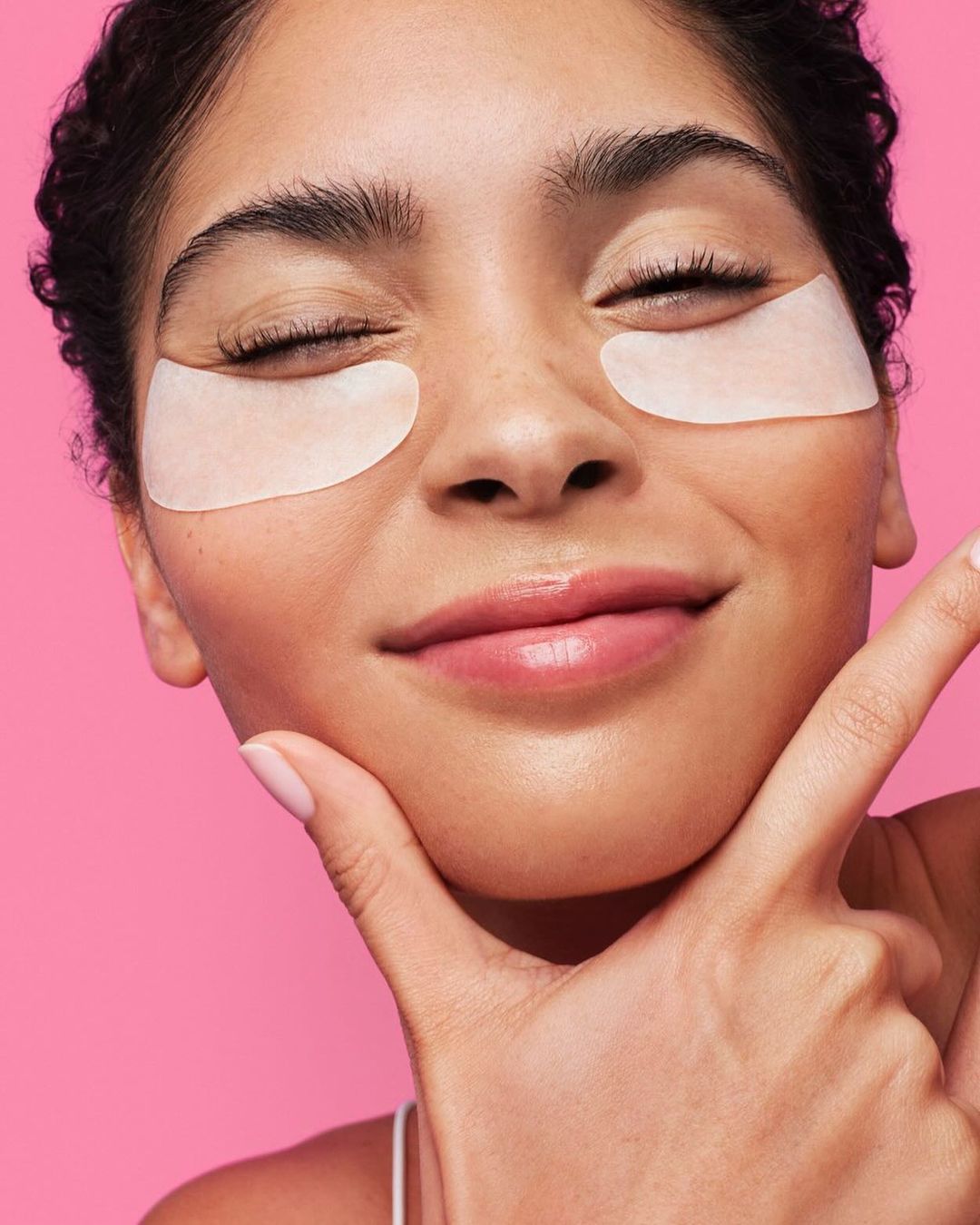 Close-up of a smiling adult female with under-eye patches, fair skin, and dark hair against a pink background, beauty concept.