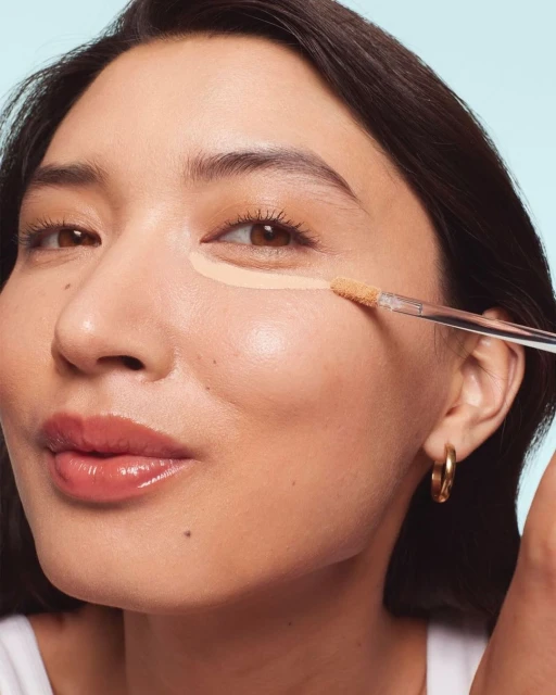 Close-up of a woman applying concealer under her eye with a makeup brush, showcasing beauty and makeup application.