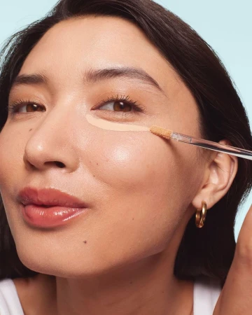 Close-up of a woman applying concealer under her eye with a makeup brush, showcasing beauty and makeup application.