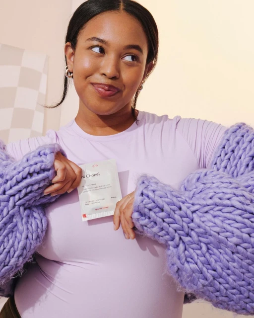 Young woman in purple sweater and top holds a cosmetic product, looking up and to the side in a studio setting with neutral backdrop.