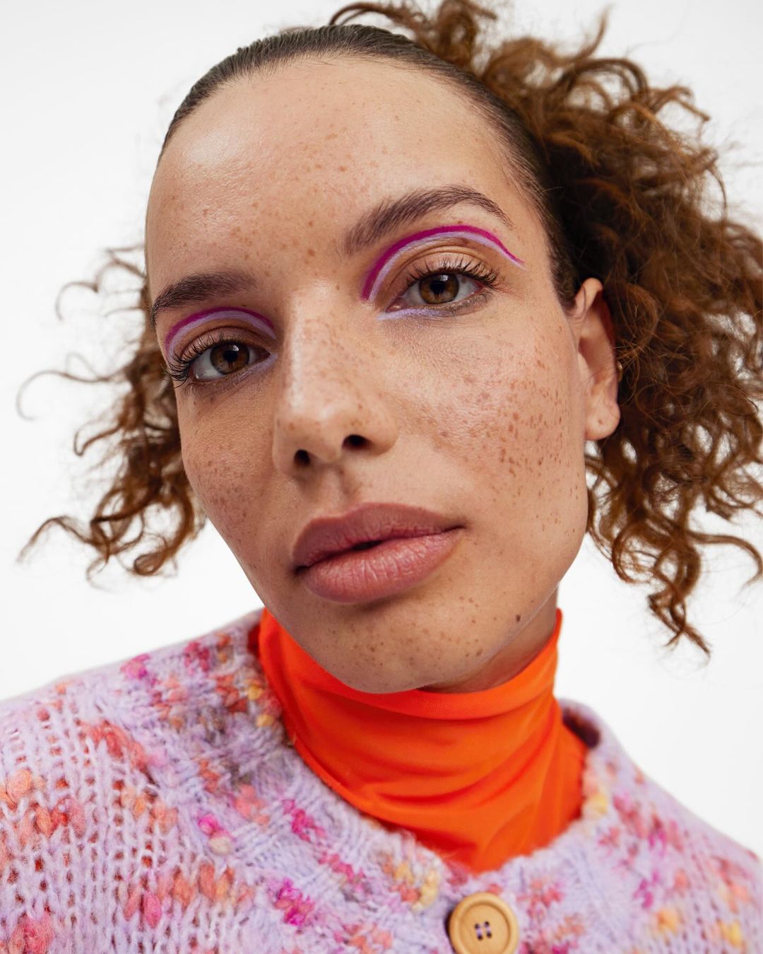 Close-up of a woman with graphic purple and pink eyeliner, fair skin with freckles, and curly brown hair, wearing a pastel cardigan.