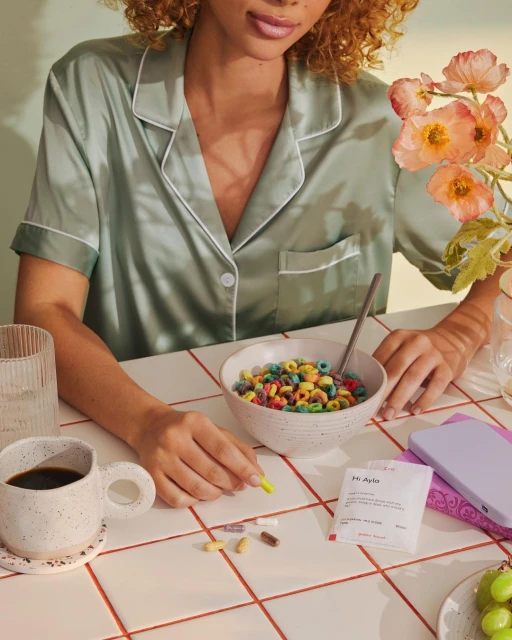 Woman in pajamas at breakfast table with colorful cereal, coffee, flowers, supplements, and phone on tiled surface in an indoor setting.