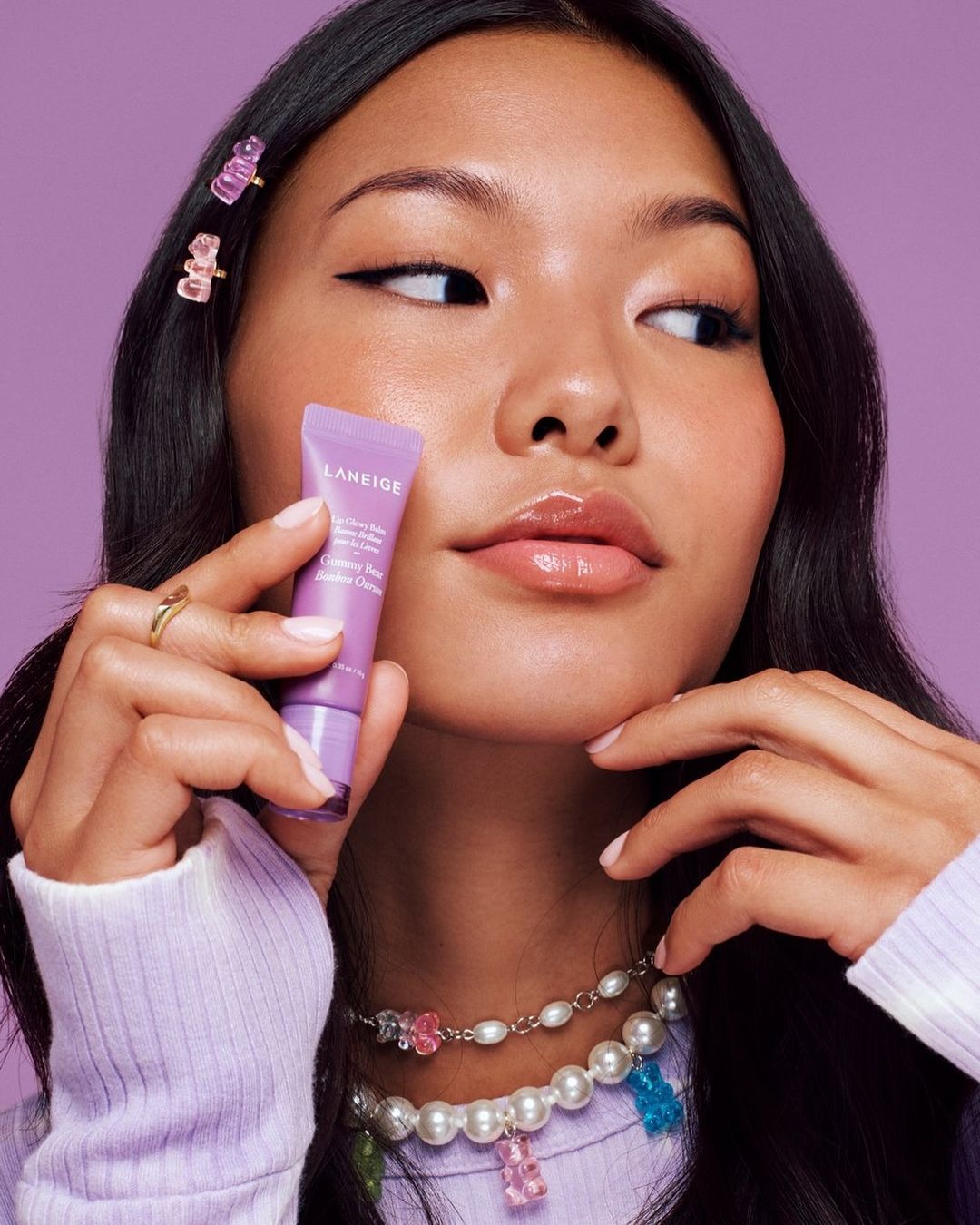 Asian woman displays a Laneige lip product with gummy bear hair clips and necklace against a lilac backdrop in a beauty product shot.
